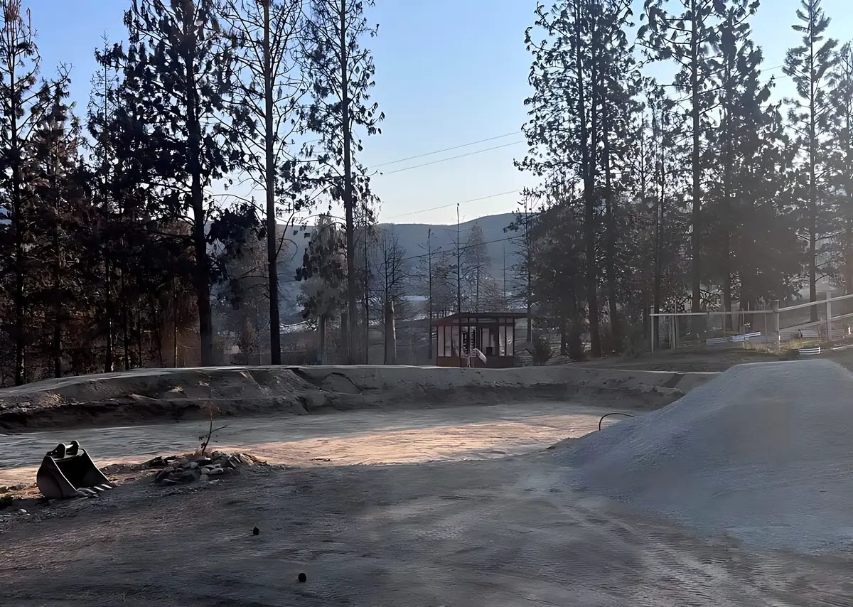Freshly graded pad with a pile of crushed rock and an excavator bucket at the edge, pines behind