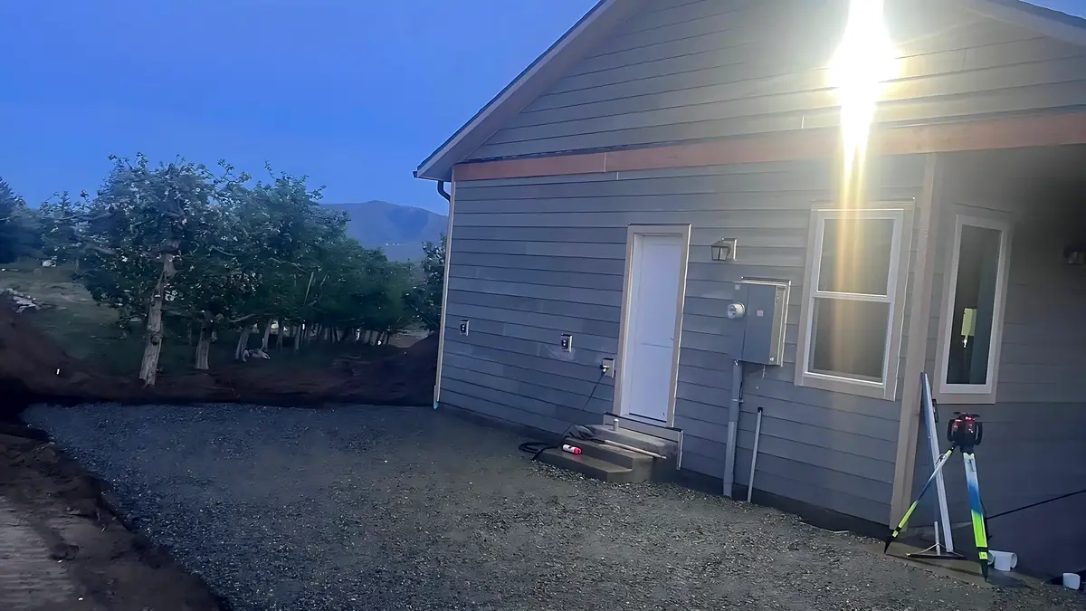 Crushed rock pad beside a new house. Graded to a laser level along the foundation, orchard behind.